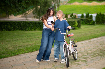 Fototapeta premium Teen boy showing something on smartphone to mom, both laughing near bicycle in park