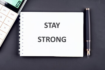 A white spiral notebook displays STAY STRONG in bold letters, next to a fountain pen and calculator on a dark grey surface