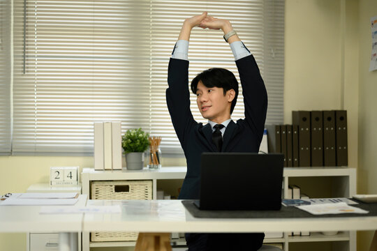 An Asian male office worker in a suit stretches his arms to relieve tension after working long hours at his desk