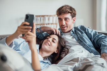 A young couple relaxing on a cozy sofa at home, smiling together while the woman uses her smartphone. Warm light, love, and comfort create a natural modern lifestyle scene.
