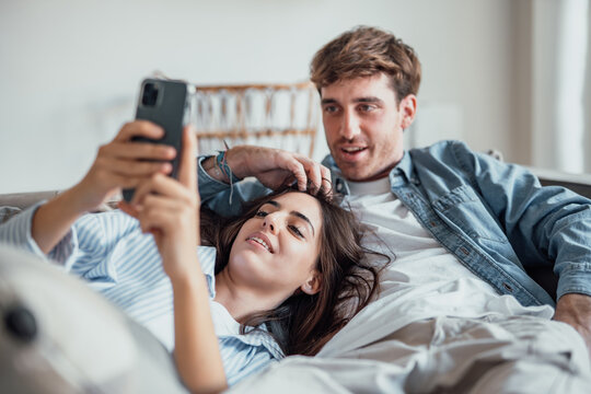 A young couple relaxing on a cozy sofa at home, smiling together while the woman uses her smartphone. Warm light, love, and comfort create a natural modern lifestyle scene.