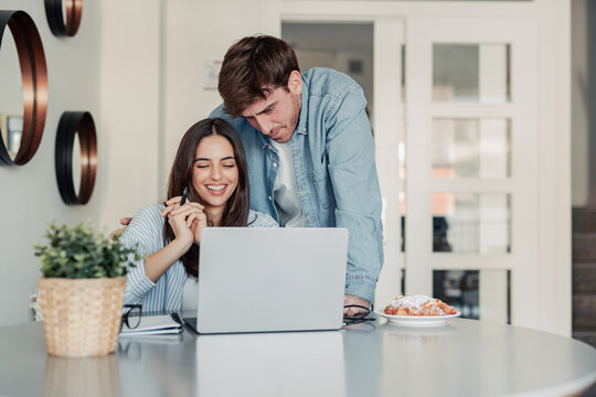 A young couple enjoys a morning at home, smiling while working together on a laptop. The warm light and teamwork reflect love, productivity, and a modern lifestyle balance.