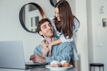 A smiling woman stands behind her partner as he works on a laptop at home. Their teamwork and shared focus reflect motivation, love, and the balance of modern relationship goals.