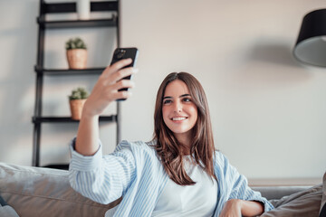 Happy pleasant millennial woman relaxing on comfortable couch, holding smartphone in hands. Smiling...