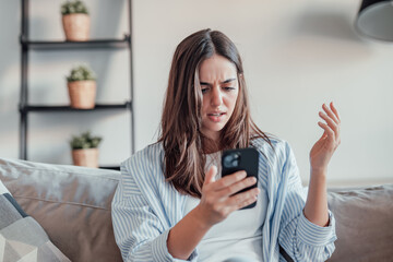 A young woman sits on the sofa, frustrated and upset while using her smartphone. Slow connection, spam, and technical issues leave her stressed and confused at home.