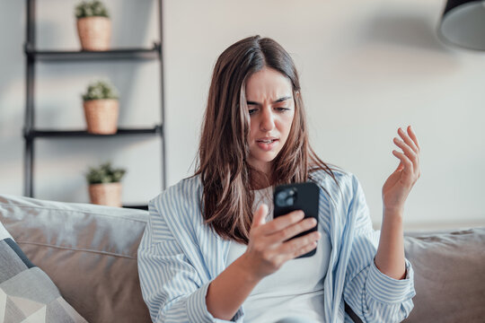 A young woman sits on the sofa, frustrated and upset while using her smartphone. Slow connection, spam, and technical issues leave her stressed and confused at home.