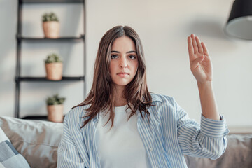 A young woman sits in her living room, showing the feminist gesture with a sad and concerned...