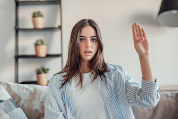 A young woman sits in her living room, showing the feminist gesture with a sad and concerned...