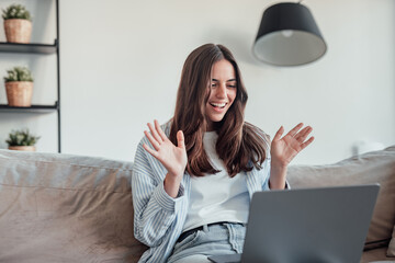 Smiling girl sit on couch at home wave talk with friend or family having video call on laptop,...