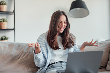 Smiling girl sit on couch at home wave talk with friend or family having video call on laptop,...