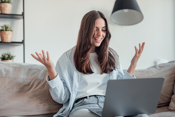 Smiling girl sit on couch at home wave talk with friend or family having video call on laptop,...