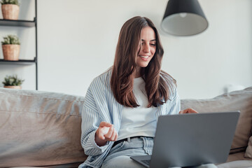 Smiling girl sit on couch at home wave talk with friend or family having video call on laptop,...