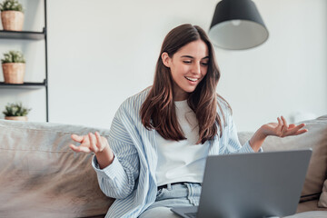Smiling girl sit on couch at home wave talk with friend or family having video call on laptop,...