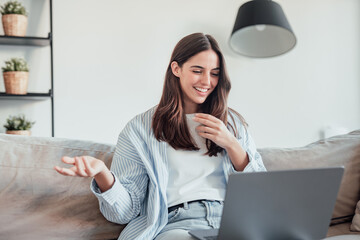 Smiling girl sit on couch at home wave talk with friend or family having video call on laptop,...
