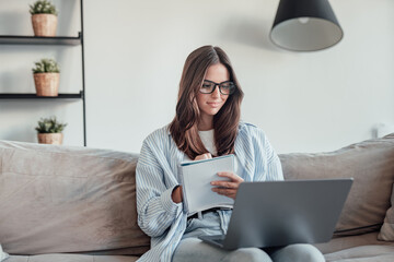 A young woman sits on the sofa wearing glasses, writing in a notebook while using her laptop. She...