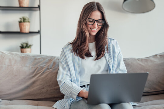 A young woman sits on the sofa wearing glasses, focused on her smartphone. She works, browses, and shops online at home, reflecting concentration, professionalism, and a modern freelancer lifestyle.