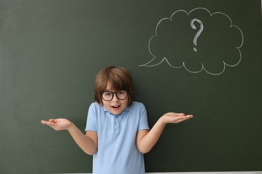 Back to school. Confused boy near chalkboard with drawn thought bubble and question mark indoors