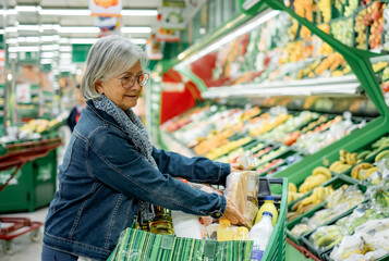 Smiling senior woman pushing a shopping cart in the supermarket in the fruit and vegetable department shopping with attention to quality and prices - Concept of consumerism, price rise, inflation
