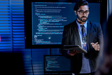 An IT support male engineer dressed smartly with a headset actively assists a user analyzing system data on his computer in a dimly lit control room setting ensuring smooth operations.