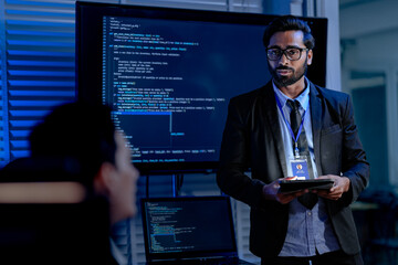An IT support male engineer dressed smartly with a headset actively assists a user analyzing system data on his computer in a dimly lit control room setting ensuring smooth operations.
