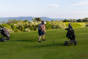 Teeing off, Female golfer preparing to hit the ball on golf course in rural Shropshire. Her golf bag and trolley are near by with a selection of clubs ready for use.