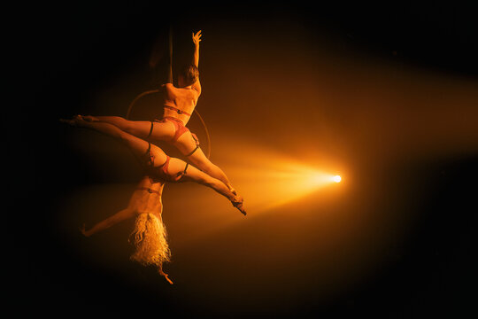 Two female aerialists on hoop in an inverted duo split under golden spotlight—strength and flexibility; horizontal with wide right-side negative space.