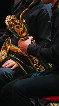 A close-up of two musicians seated on stage. One of them plays a shiny brass baritone saxophone. The orchestra performs
