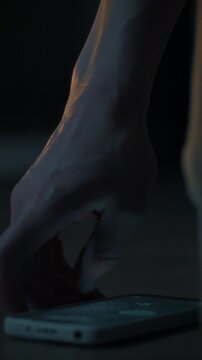 A man's hand extends toward a smartphone that glows brightly on the floor beside a bed. The scene captures a moment in the stillness of night within a bedroom setting