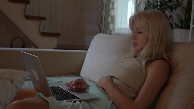 A young woman with blond hair sits on the sofa and looks at the laptop.