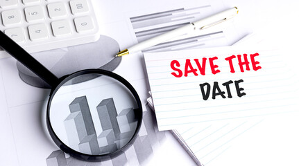 SAVE THE DATE message on notecard with magnifying glass, pen, calculator and business charts in office setting