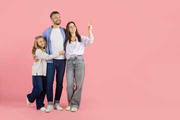 Fototapeta premium Family standing and looking same direction in studio. Daughter hugging father, mother pointing upward, joyful and optimistic concept of family vision and future goals