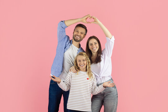 Happy young family posing in studio on pink background, smiling together. Cheerful parents and daughter showing heart symbol with hands, expressing love and unity - Powered by Adobe