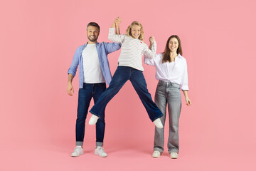 Obraz premium Happy parents lifting daughter in air in studio, playful family moment against pink backdrop. Smiling faces expressing joy, support, and strong emotional connection