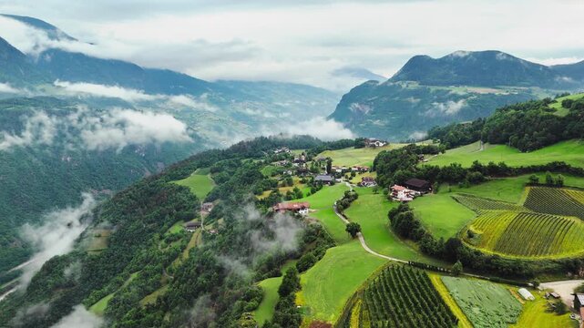 Patchwork meadows and hillside farms across a Dolomite valley, aerial view. Overcast conditions with cloud bands at mid-elevation