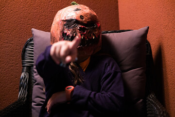 Young person wearing a scary Halloween pumpkin mask, pointing at the camera with a finger, sitting in a chair on a terrace. Fear, Jack-o'-Lantern, celebration, terror, and carnival.