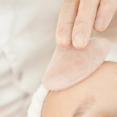 Young caucasian female receiving facial massage with rose quartz gua sha stone in spa treatment.