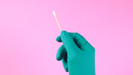 A hand wearing a green medical glove holding a cotton swab on pink backgroun