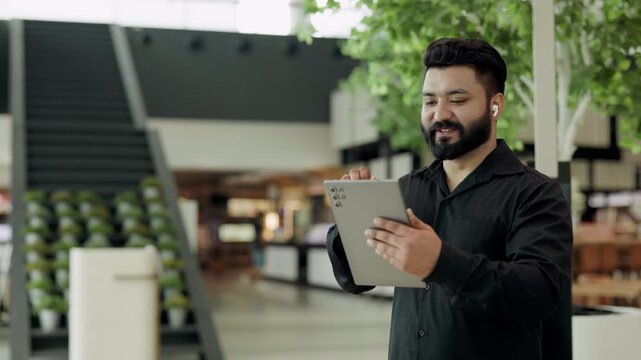 Portrait of friendly Indian salesman with earphones waves hand to interlocutor on tablet in office. South Asian businessman joins video-call via mobile device in shopping mall
