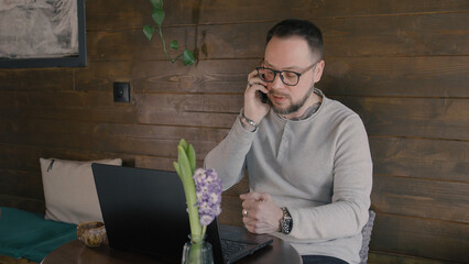 Man talking on the phone while working on a laptop in a cozy cafe with a flower vase on the table during the day