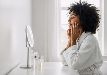 Young woman in bathrobe touching her face while looking in the mirror during skincare routine at home.