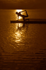 Silhouette woman practicing hatha yoga asana or stretching on the beach pier at evening sunrise Yoga lady female girl performs exercises during sunset on the sea river ocean pond lake coast Reflection