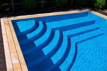 Modern pool with clear blue water and elegant geometric steps casting shadows. Sunlight reflects off the ripples, surrounded by stone edging and tree shadows