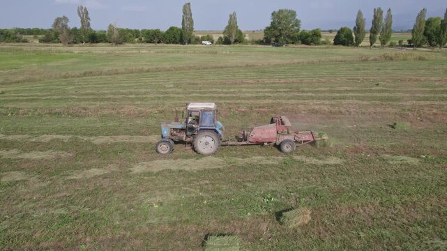 Tractor uses trailed bale machine to collect straw in field. Haymaking. Tractor collecting hay and making hay bales. Haystack making machine. Tractor Working on Farm at Sunset Time.