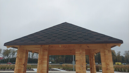 Wooden gazebo with a hexagonal roof stands against a cloudy sky in an open area near a highway