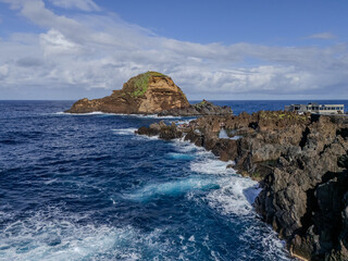 Natural volcanic swimming pools surrounded by rugged lava rocks in Porto Moniz, Madeira, with a scenic ocean backdrop and a small island offshore. 

