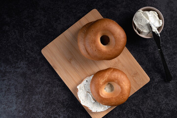 A high angel view of Two bagels with cream cheese on top of a wooden cutting board. The bagels are cut in half and the cream cheese is spread on top