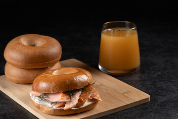 A bagel with atlantic salmon and a glass of juice are on a wooden board. The sandwich is a smoked salmon bagel with cream cheese and dill. Two plain bagels on the side