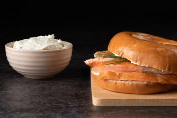 A sandwich with salmon and dill is on a wooden cutting board. A bowl of cream cheese is next to the sandwich