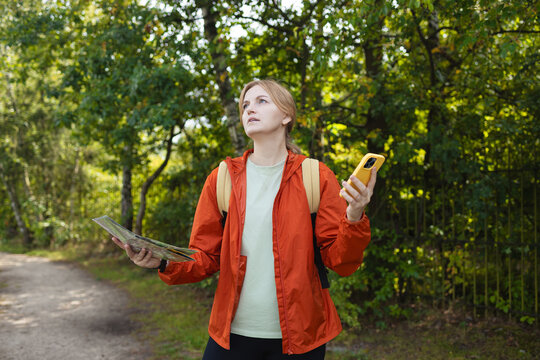 Young woman traveler wearing orange jacket holding map and smartphone looking around while hiking in green forest. Concept of navigation error, lost signal, outdoor adventure, and travel difficulties.