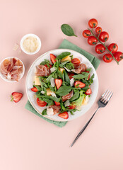 Colorful Italian arugula salad with prosciutto, strawberries, melon, cherry tomatoes, cheese on pink background.Top view